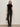A woman with straight brown hair and a neutral expression stands against a plain wall wearing the Sanctuary Clothing slick brook bootcut pant in black, paired with a black sequin top, black boots, and holding a leopard print handbag.