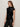 A woman with long straight brown hair stands sideways against a plain background, wearing the Sanctuary Clothing funnel neck sequin top in black with black pants and a neutral expression.