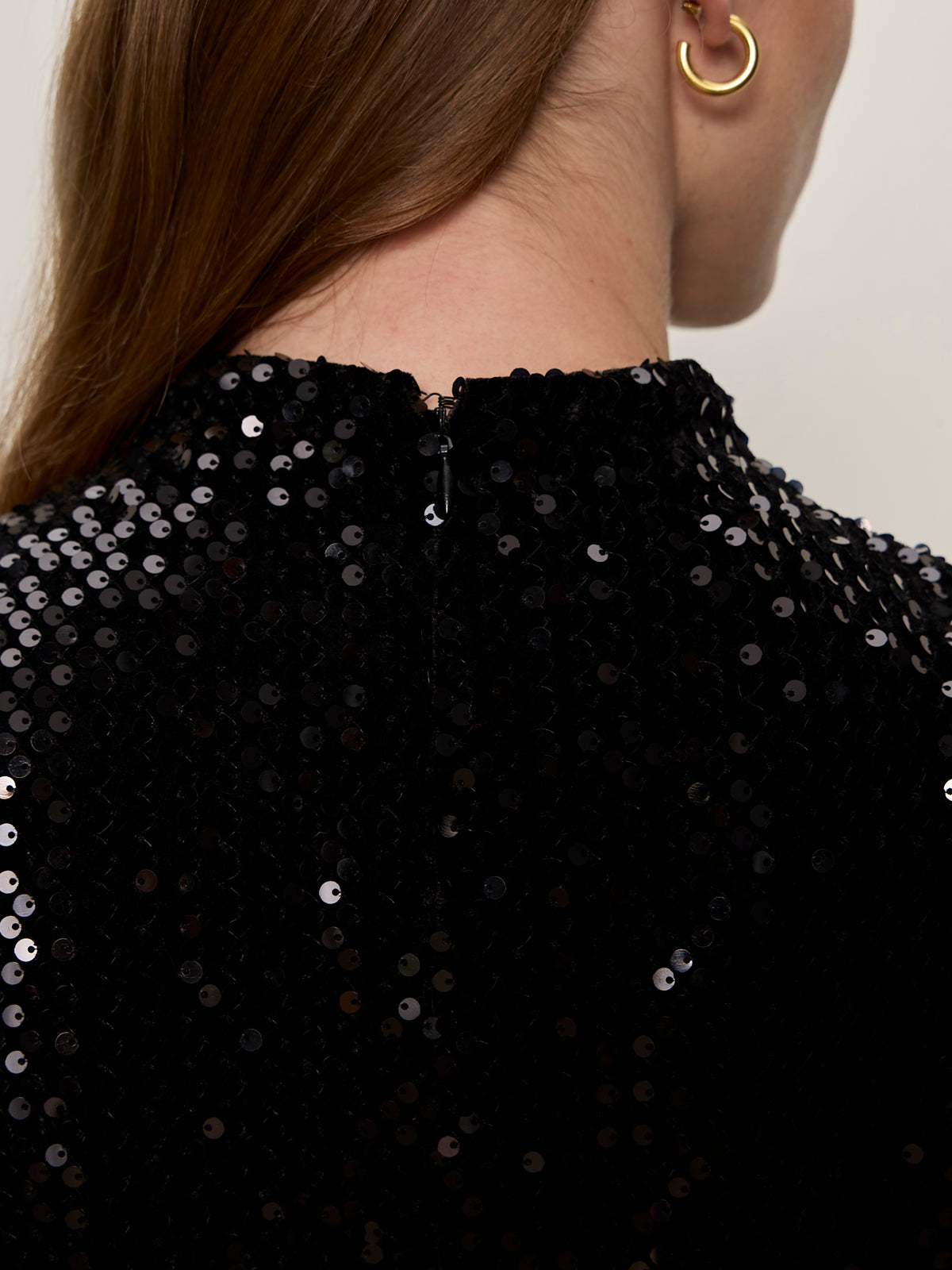 Close-up of a person’s upper back in the Sanctuary Clothing funnel neck sequin top in black, featuring a small back zipper. The person has long brown hair and wears a gold hoop earring.