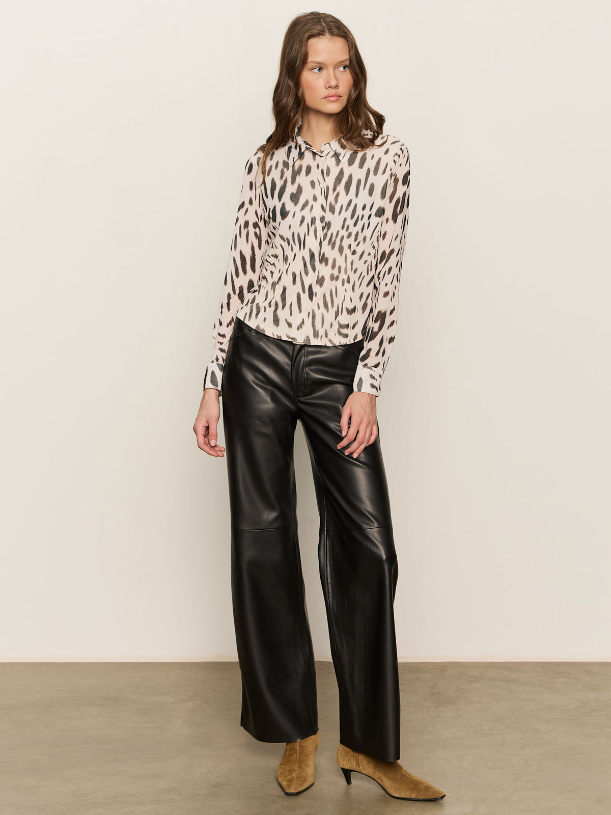 A woman with wavy brown hair and a neutral expression wears the Sanctuary Clothing fitted mesh button down top mother leo—a long-sleeved animal print blouse—against a plain background. She pairs it with black wide-leg leather pants and tan pointed-toe ankle boots.