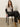 A woman sits on a black chair wearing the Sanctuary Clothing fitted mesh button down top mother leo, paired with black leather pants and tan suede boots, against a plain light background.