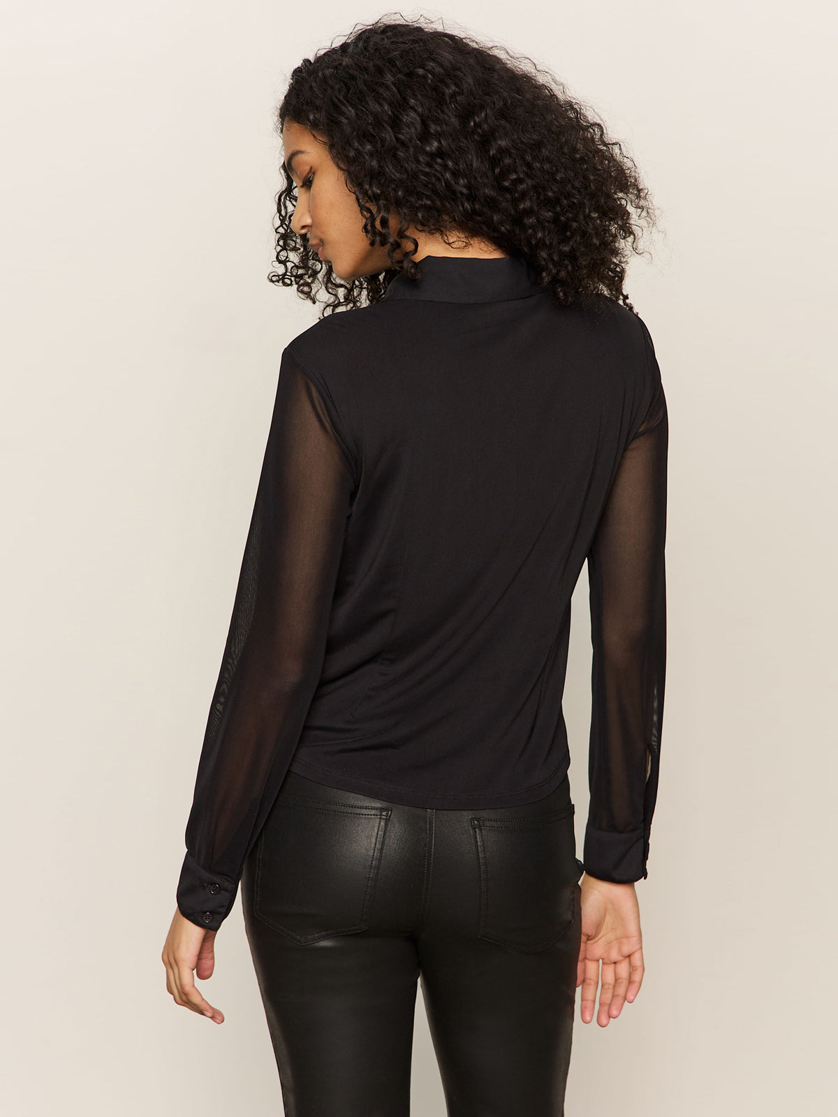 A woman with curly hair is seen from the back, wearing the Sanctuary Clothing fitted mesh button down top in black and black leather pants, standing against a plain light background.