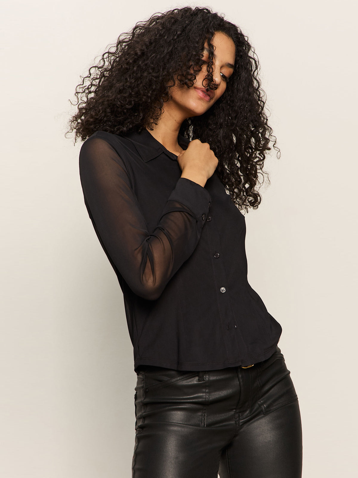 A woman with curly hair wears the Sanctuary Clothing fitted mesh button down top in black and black leather pants, standing against a plain light background.