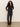A woman with long curly hair stands against a plain wall, wearing Sanctuary Clothing's fitted mesh button down top in black, paired with black leather pants and tan pointed-toe shoes, her hands resting in her pockets.