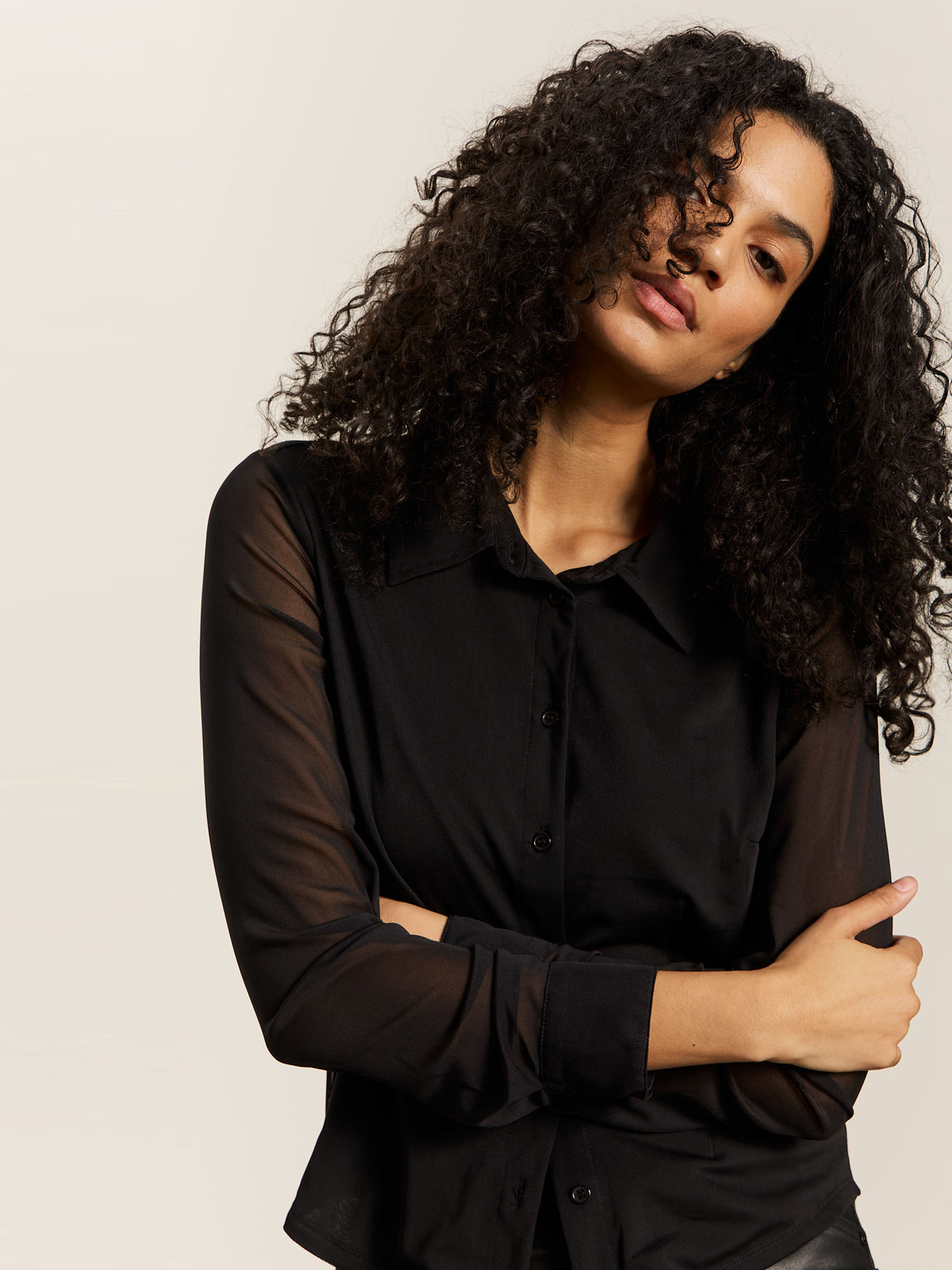 A woman with curly hair models the Sanctuary Clothing fitted mesh button down top in black, standing against a light neutral background with her arms crossed and head slightly tilted as she looks at the camera.