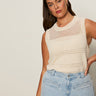 A woman with long brown hair wears the Sanctuary Clothing "waves of summer shell sweater macrame" in extended sizing, paired with light blue high-waisted jeans, standing against a plain light background with a neutral expression.