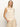 A woman stands confidently against an off-white background wearing the Sanctuary Clothing “waves of summer shell sweater macrame”—a sleeveless, light beige knit top with horizontal eyelet stripes—paired with light-colored pants.