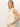 A woman with long blonde hair wears the Sanctuary Clothing "waves of summer shell sweater macrame," a sleeveless, cream knit top with horizontal patterns, paired with light beige pants, standing against a plain light background.