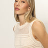 A woman with long blonde hair wears the Sanctuary Clothing "Waves of Summer Shell Sweater Macrame," a sleeveless, cream knit top. She looks slightly to the side with a neutral expression against a plain, light background.
