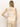 A woman with long, straight blonde hair is seen from the back, wearing Sanctuary Clothing's Waves of Summer Shell Sweater Macrame, a sleeveless, cream-colored knit top, paired with light beige pants against a plain light background.
