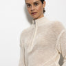 A dark-haired person wears the Sanctuary Clothing's "open knit 1/2 zip sweater eco natural," a light beige knitted sweater with a zipper, against a white background. The sweater features a textured, open-knit design as they face the camera.