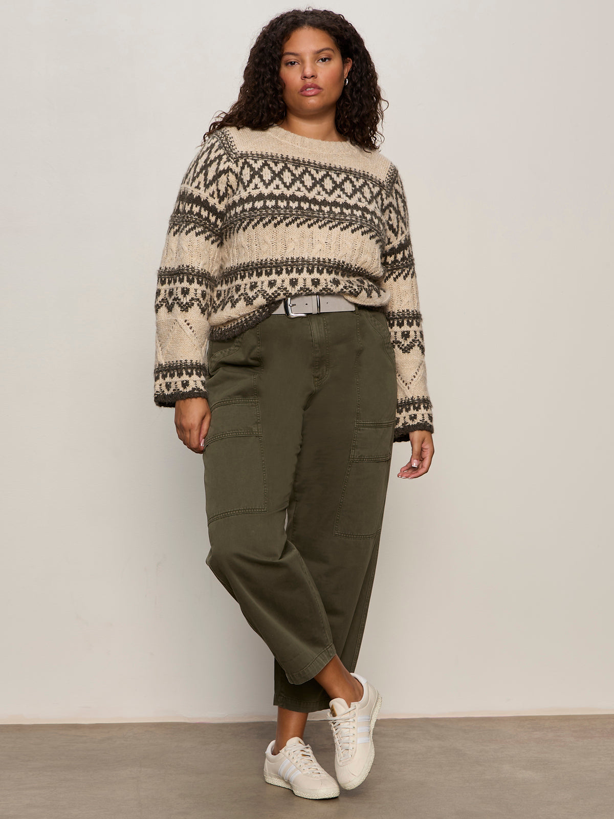 A woman with curly hair and a relaxed expression stands against a plain wall, wearing the Sanctuary Clothing fairisle sweater in light oat multi (extended sizing), olive green wide-leg cargo pants, white sneakers, and a light-colored belt.