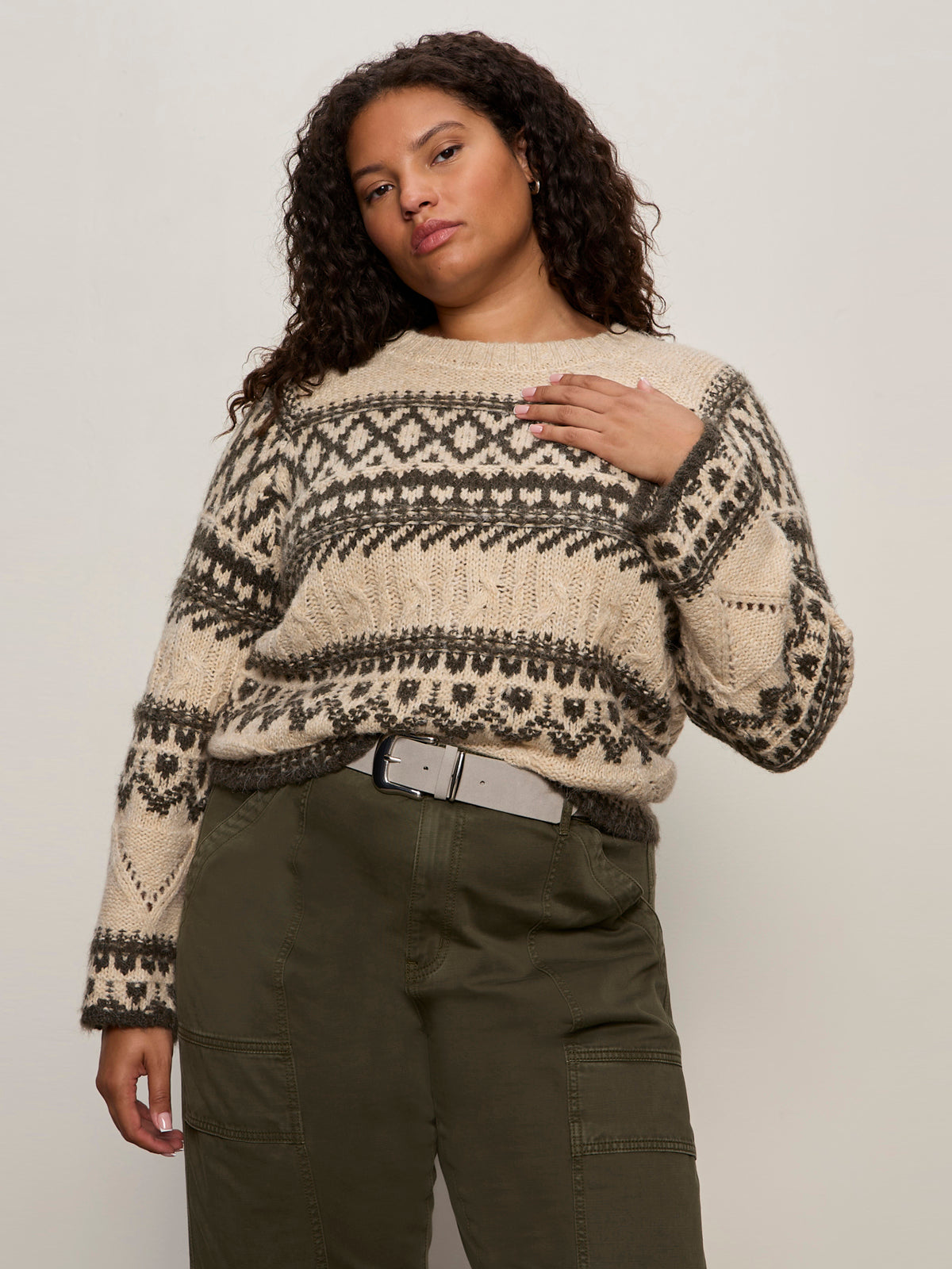 A woman with curly hair wears the Sanctuary Clothing fairisle sweater in light oat multi (extended sizing), tucked into high-waisted olive green pants with a belt, standing against a plain light background.