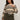A woman with curly hair wears the Sanctuary Clothing fairisle sweater in light oat multi (extended sizing), tucked into high-waisted olive green pants with a belt, standing against a plain light background.