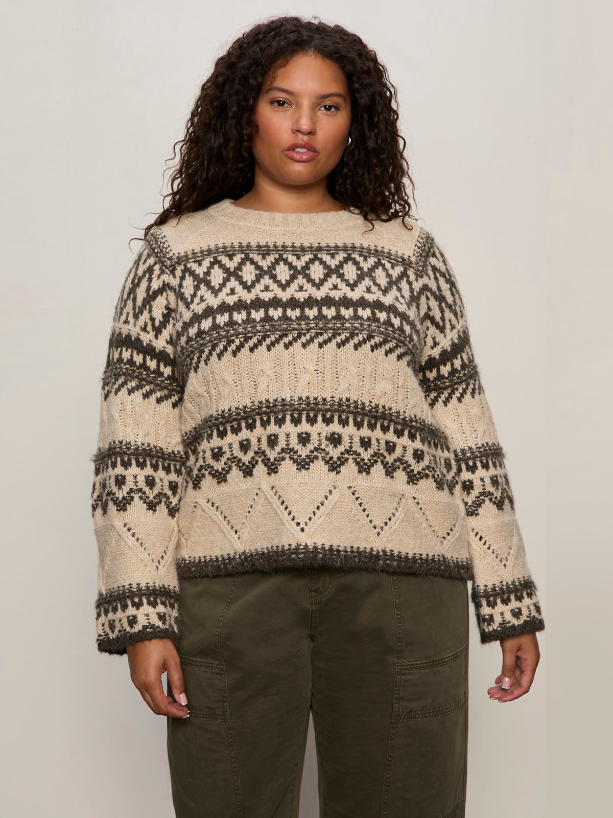 A woman with curly hair wears the Sanctuary Clothing fairisle sweater in light oat multi (extended sizing), paired with olive green pants, standing against a plain light background.