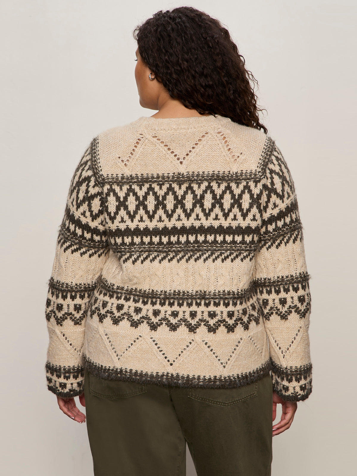 A person with curly hair is seen from the back wearing Sanctuary Clothing’s fairisle sweater light oat multi (extended sizing) with black geometric patterns and olive green pants.