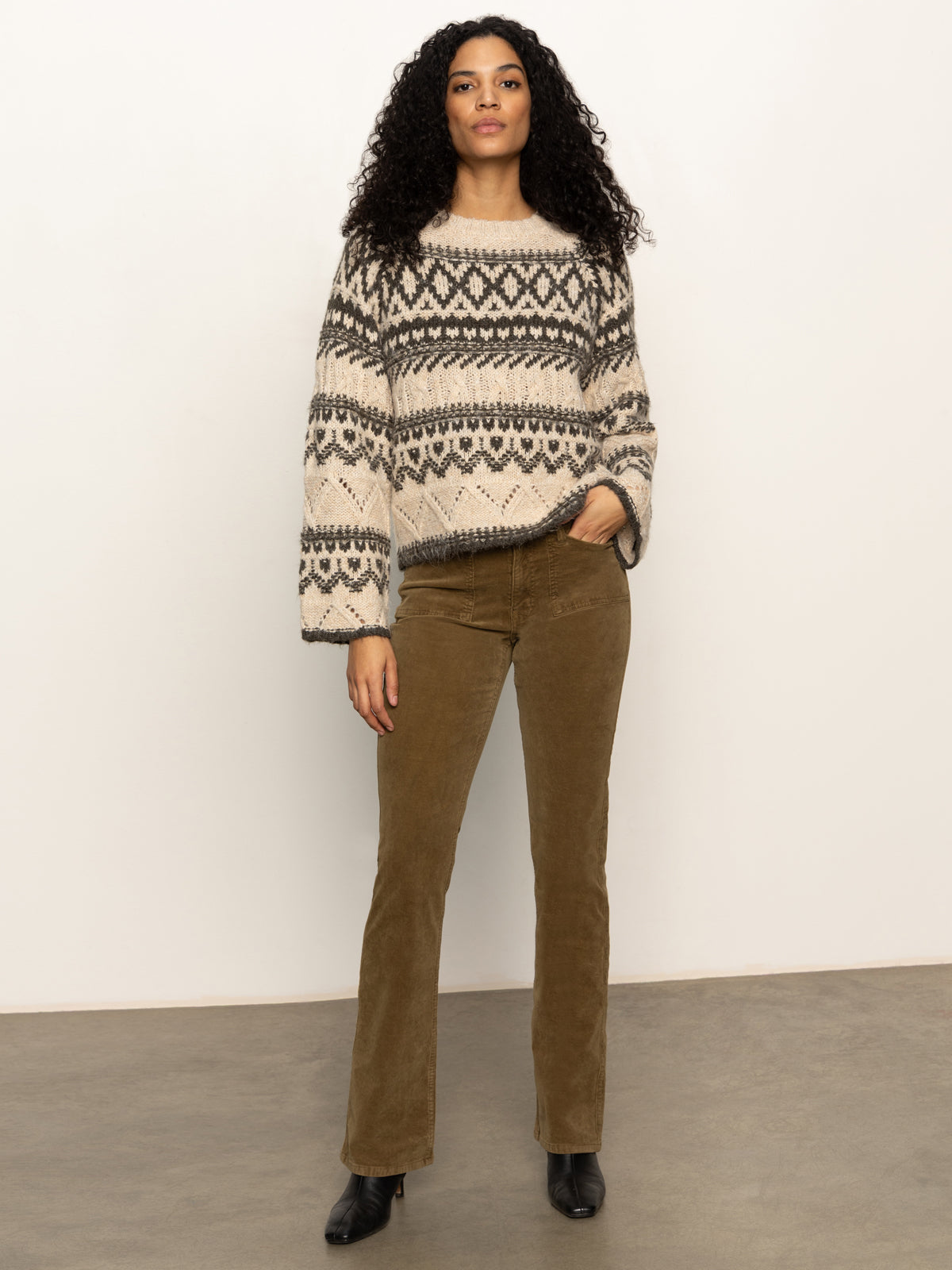A woman with curly hair wears the Sanctuary Clothing fairisle crew neck sweater in light oat multi, paired with brown corduroy pants and black boots, standing against a plain light-colored wall.