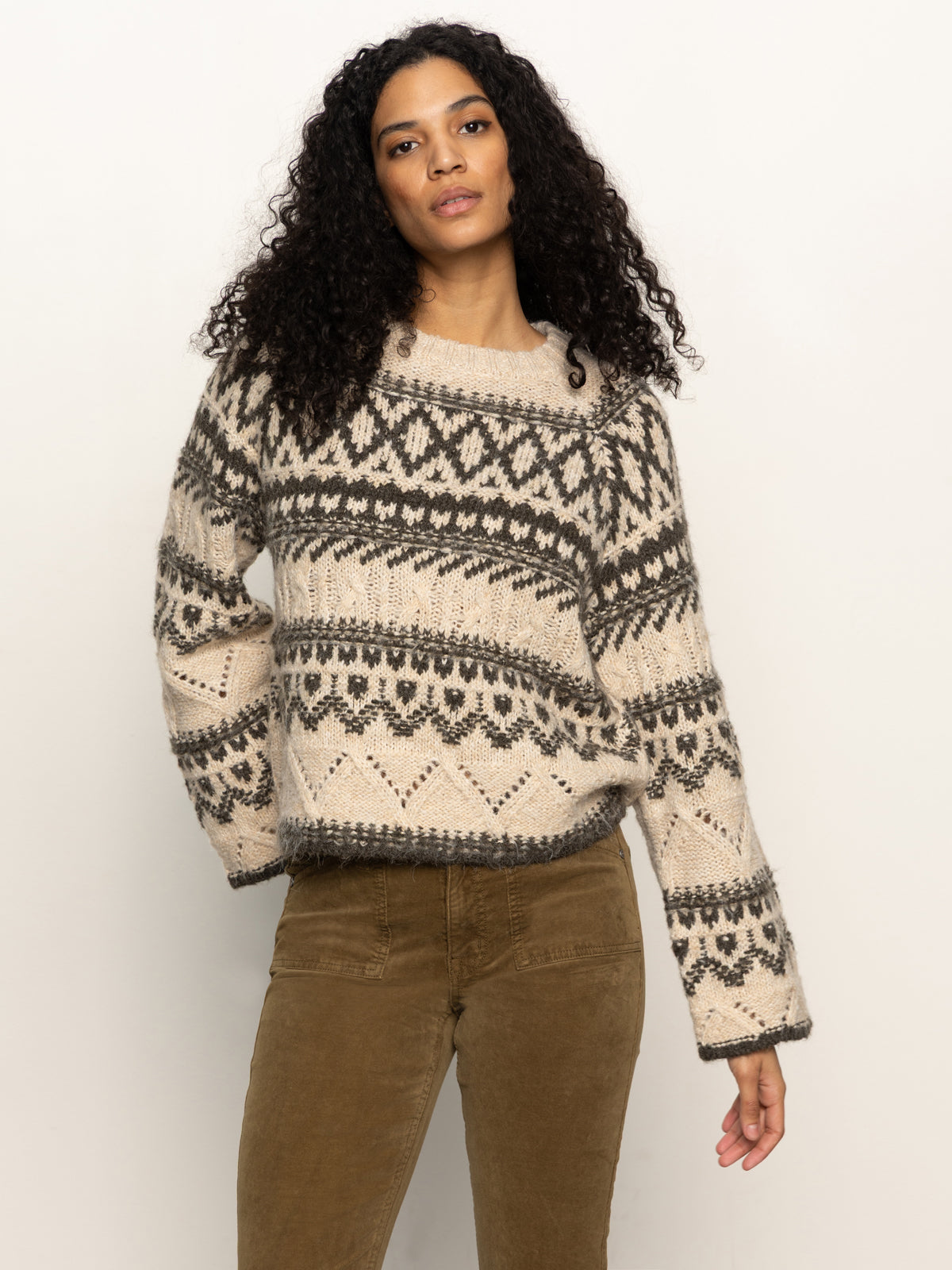 A woman with curly dark hair wears the Sanctuary Clothing fairisle crew neck sweater in light oat multi and brown corduroy pants, standing against a plain off-white background.