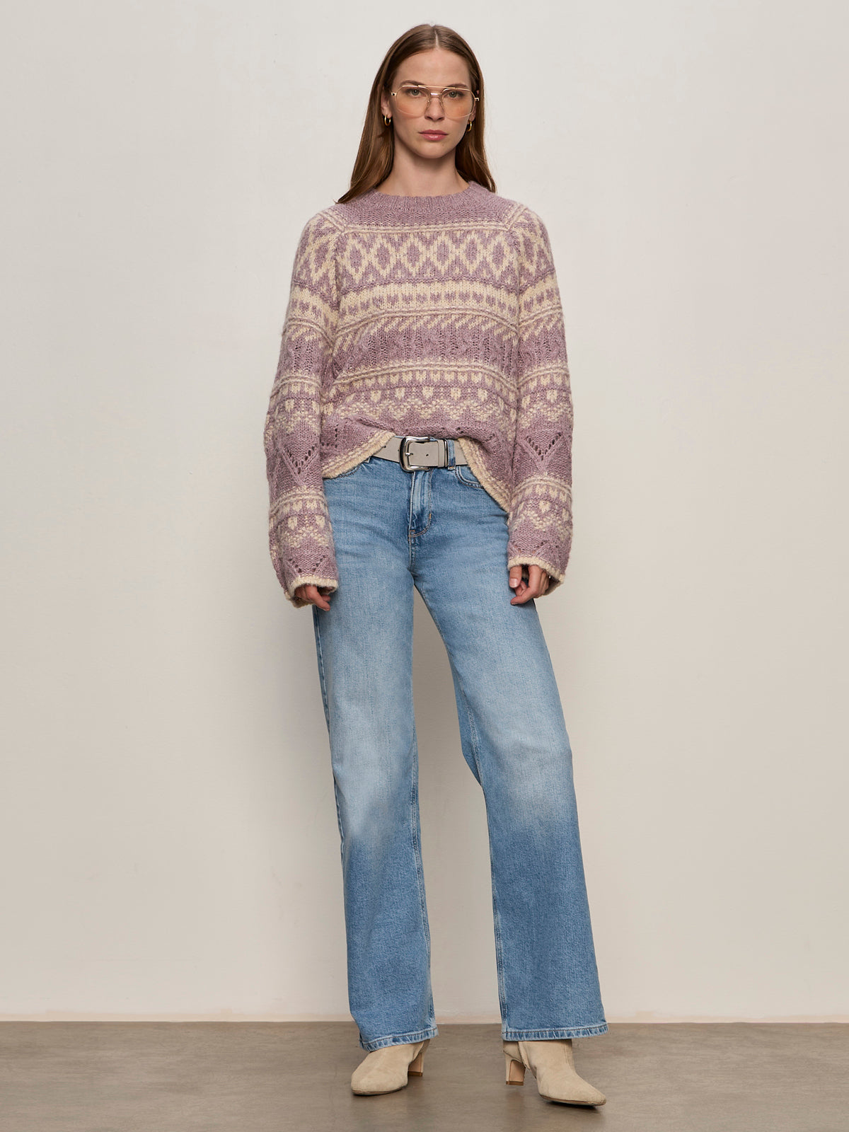 A woman stands against a light background wearing glasses and the Sanctuary Clothing fairisle crew neck sweater in twilight mauve multi, paired with light blue high-waisted jeans, a gray belt, and beige heeled boots.