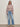 A woman stands against a light background wearing glasses and the Sanctuary Clothing fairisle crew neck sweater in twilight mauve multi, paired with light blue high-waisted jeans, a gray belt, and beige heeled boots.