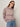 A person wearing the Sanctuary Clothing fairisle crew neck sweater in twilight mauve multi stands against a plain off-white background, paired with tinted sunglasses, light blue jeans, and a gray belt.