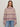 A person with straight, light brown hair wears clear glasses, the Sanctuary Clothing fairisle crew neck sweater in twilight mauve multi, and light blue jeans, standing against a plain light background.