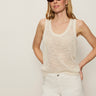 A woman stands confidently against a plain light background, hands in pockets, wearing sunglasses, white shorts, and the Sanctuary Clothing mesh stitch shell sweater macrame sleeveless cream knit top.