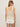 A woman with straight, light brown hair models the Sanctuary Clothing mesh stitch shell sweater macrame in cream. She pairs it with white shorts, gold hoop earrings, and sunglasses, facing away from a plain light background.