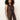 A woman with curly hair models the Sanctuary Clothing long openwork vest mesa, a sleeveless brown crochet button-up style, over light-colored pants against a plain light background.