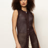 A woman with curly hair models the Sanctuary Clothing long openwork vest mesa, a sleeveless brown crochet button-up style, over light-colored pants against a plain light background.