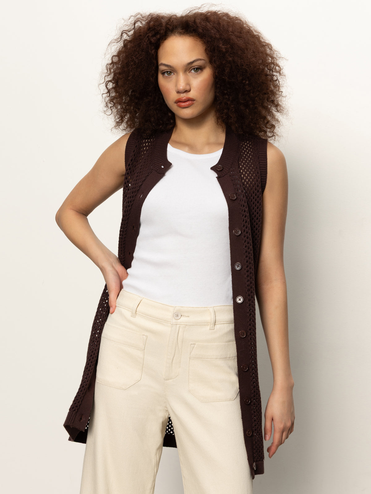 A woman with curly brown hair wears Sanctuary Clothing's long openwork vest mesa—a sleeveless, buttoned dark brown knit vest—over a white top and beige pants, standing against a plain light background with one hand on her hip.