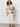 A woman stands against a plain background wearing a cream crochet short-sleeve shirt with black stripes over a black bralette, cream cropped pants, and brown sandals. She has curly hair and a relaxed pose.