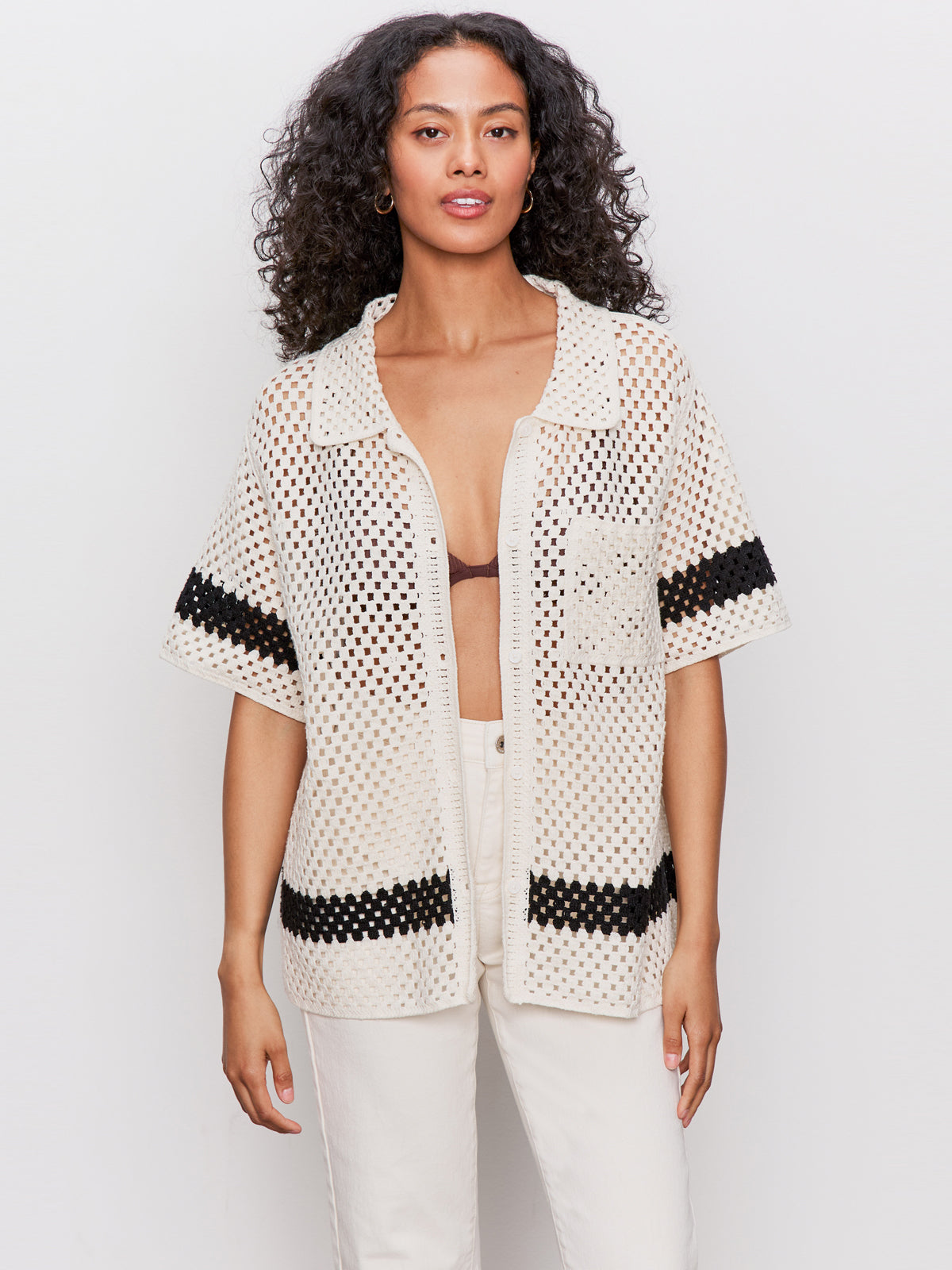 A woman with long curly hair wears a cream-colored crochet short-sleeve shirt with black stripes over a black bra, paired with off-white pants, standing against a plain light background.