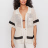A woman with long curly hair wears a cream-colored crochet short-sleeve shirt with black stripes over a black bra, paired with off-white pants, standing against a plain light background.