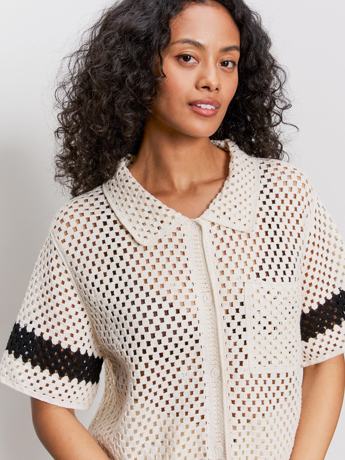 A woman with curly hair wears a cream-colored, short-sleeve, crochet top with black stripes on the sleeves and a front pocket. She stands against a plain light background, looking confidently at the camera.