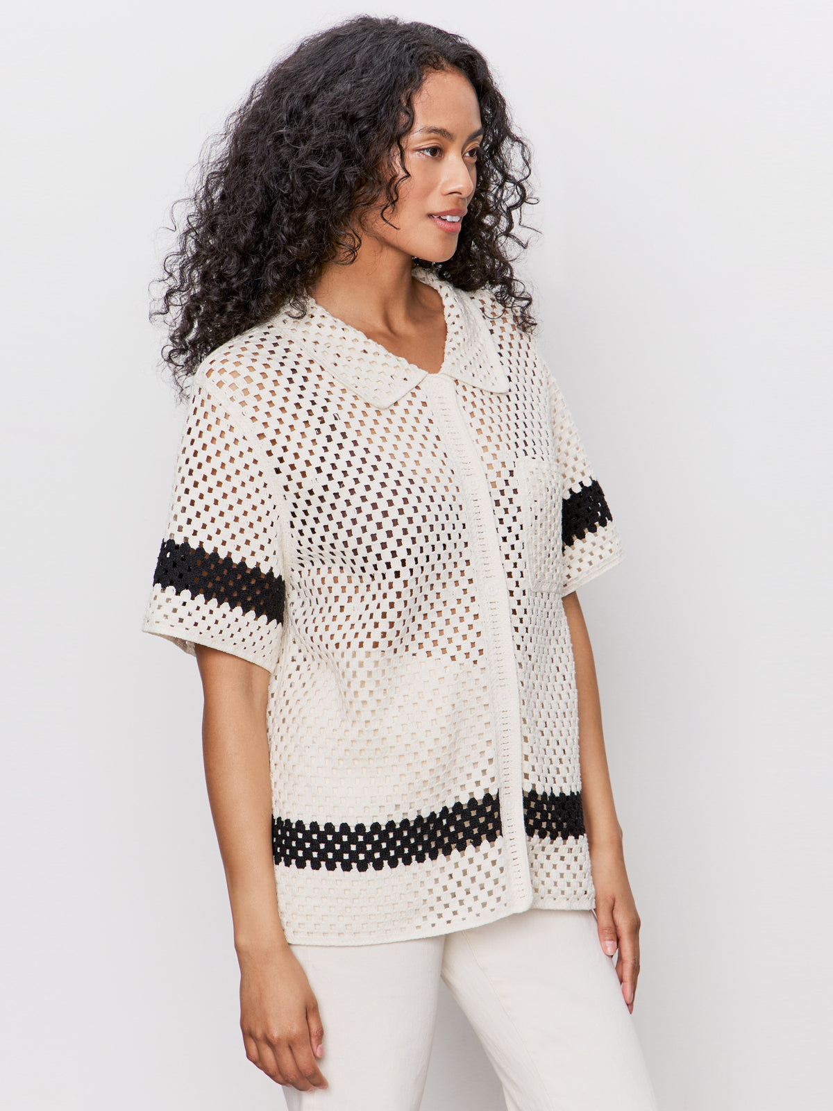 Woman with long curly hair wearing a short-sleeve, cream-colored crochet shirt with black horizontal stripes and cream pants, standing against a plain light background.