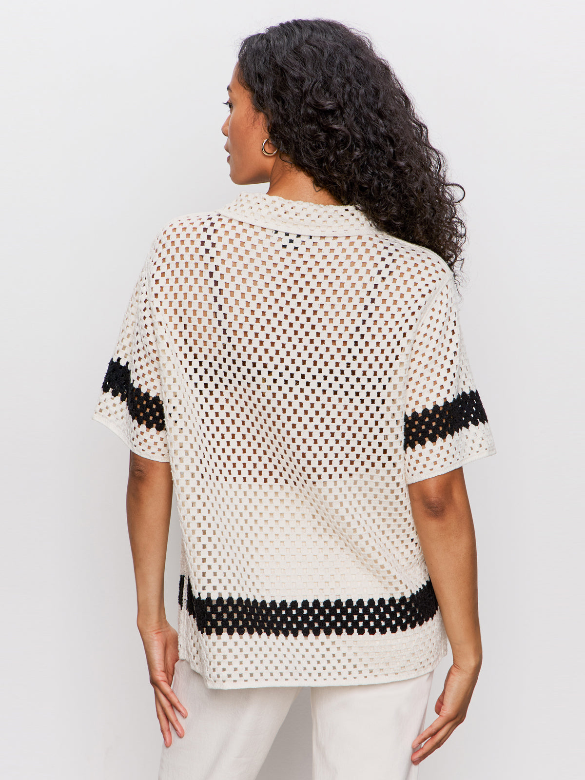 A woman with curly hair stands facing away, wearing a short-sleeve, cream-colored crochet top with black horizontal stripes on the sleeves and around the lower back, paired with white pants.