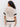 A woman with curly hair stands facing away, wearing a short-sleeve, cream-colored crochet top with black horizontal stripes on the sleeves and around the lower back, paired with white pants.