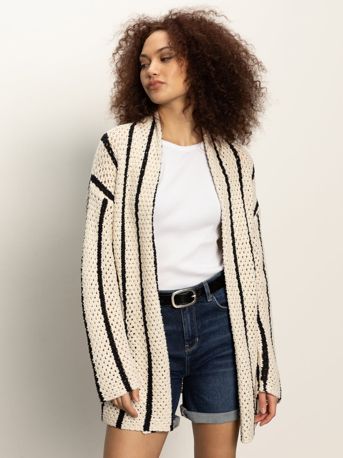 A woman with curly hair wears the Sanctuary Clothing openwork beach cardi in chalk black stripe over a white T-shirt and denim shorts, standing against a plain light background.