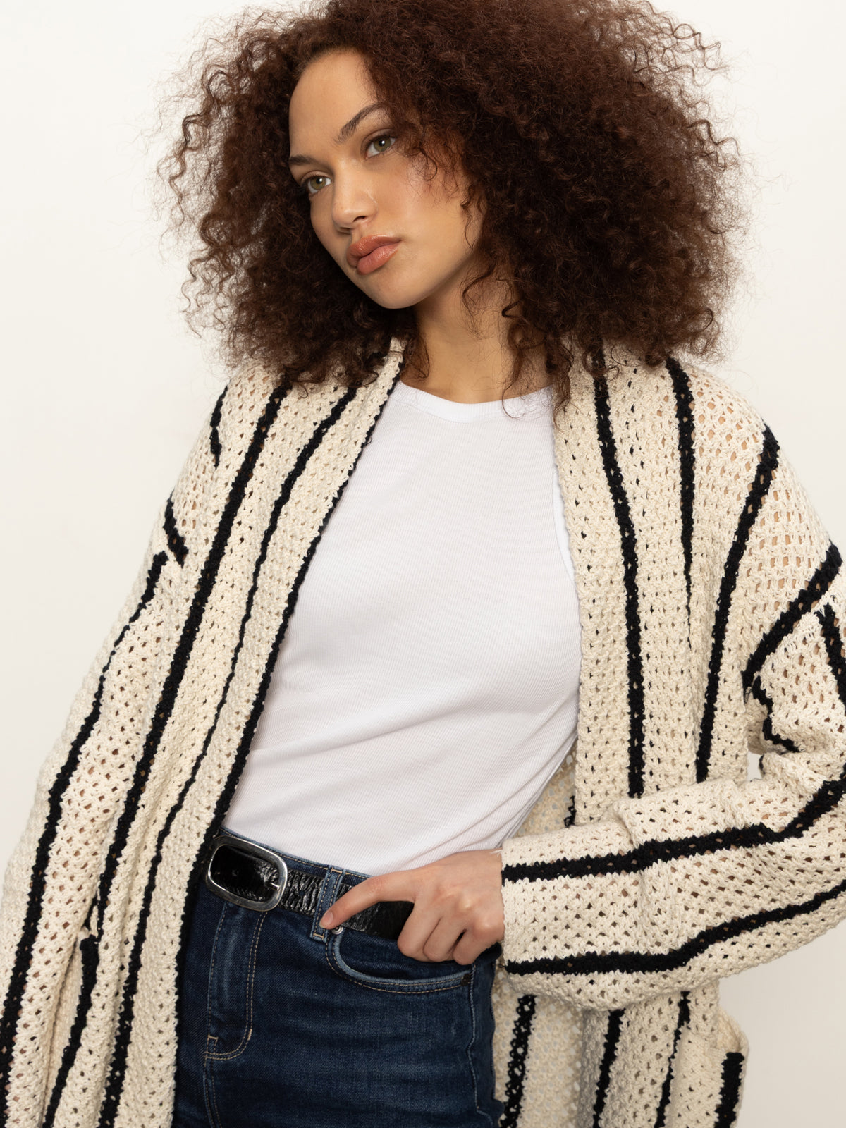 A woman with curly hair wears the Sanctuary Clothing openwork beach cardi in chalk black stripe over a white t-shirt and denim jeans, standing against a plain light background.