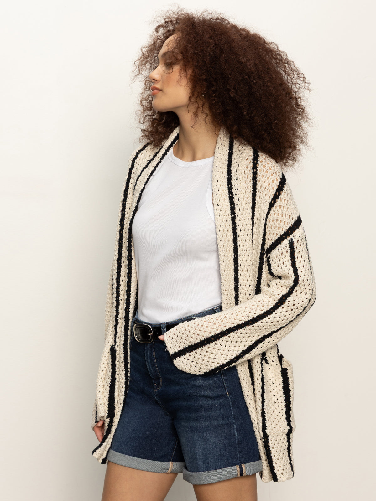 A woman with curly hair models the Sanctuary Clothing openwork beach cardi in chalk black stripe, styled over a white T-shirt and blue denim shorts, standing against a plain light background.