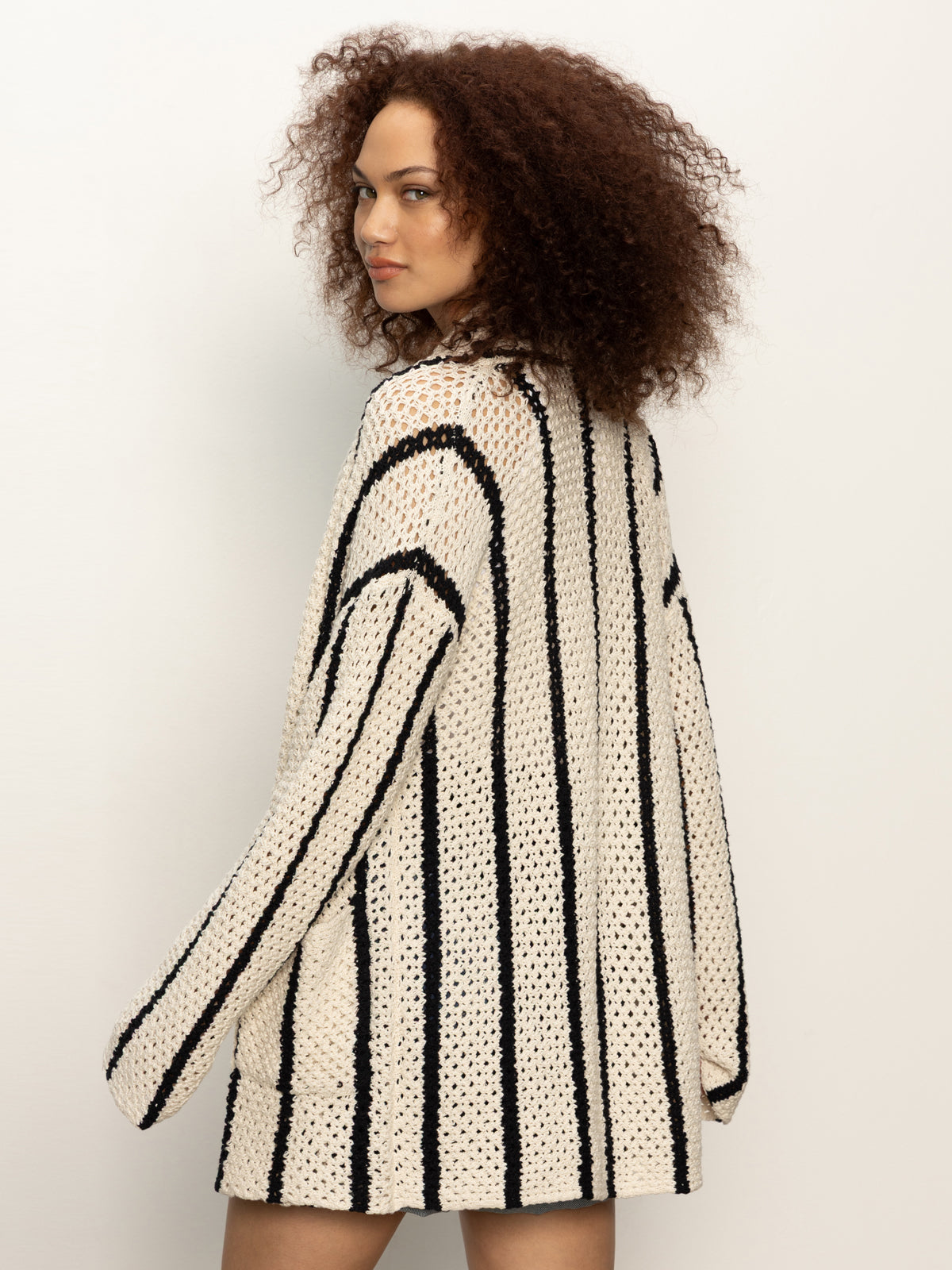 Wearing the Sanctuary Clothing openwork beach cardi in chalk black stripe, a person with curly hair looks over their shoulder against a plain light background.