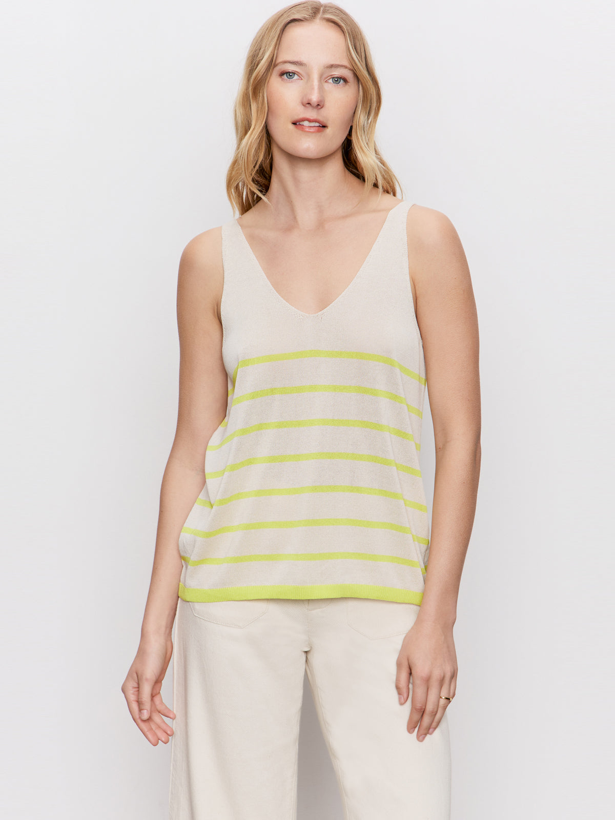A woman with blonde hair wears Sanctuary Clothing's V Neck Knitwear Tank in Muslin Desert Lime Stripe and cream pants, standing against a plain white background.