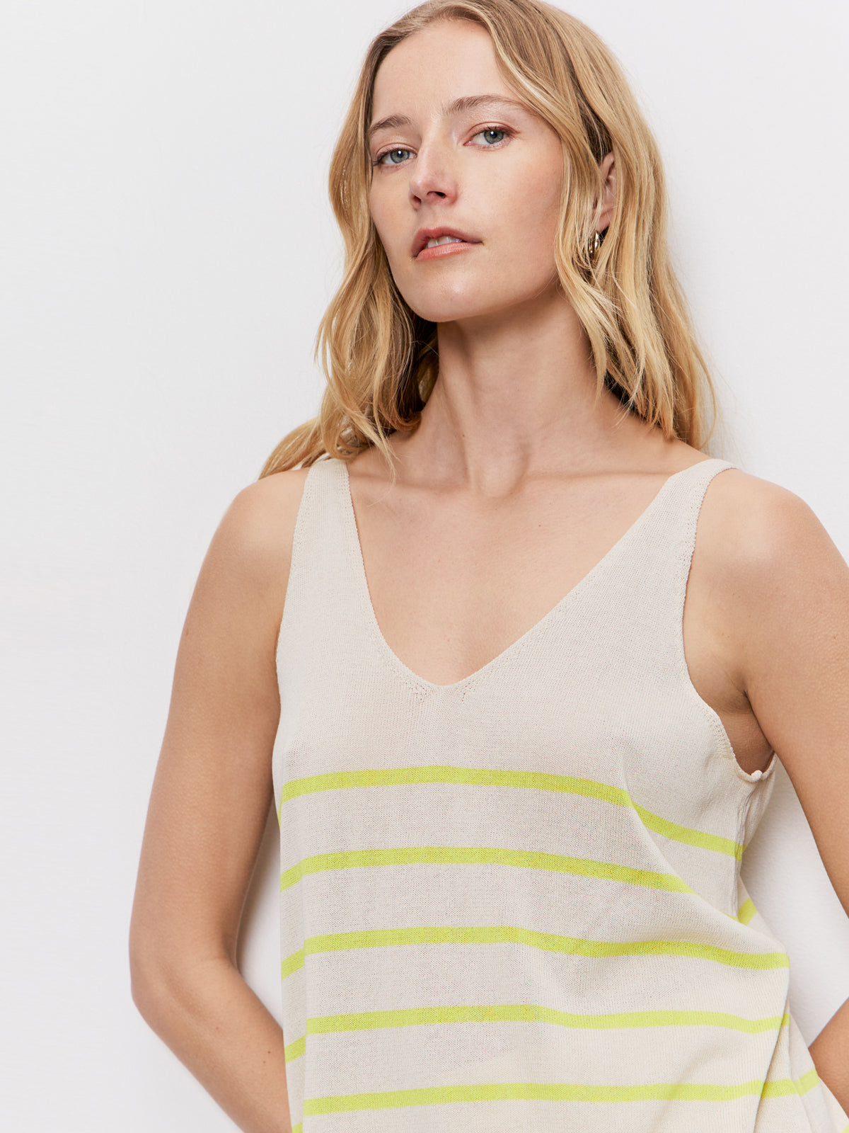 A woman with long blonde hair models the Sanctuary Clothing v neck knitwear tank in muslin desert lime stripe, a sleeveless beige top with light green horizontal stripes, against a plain white background.