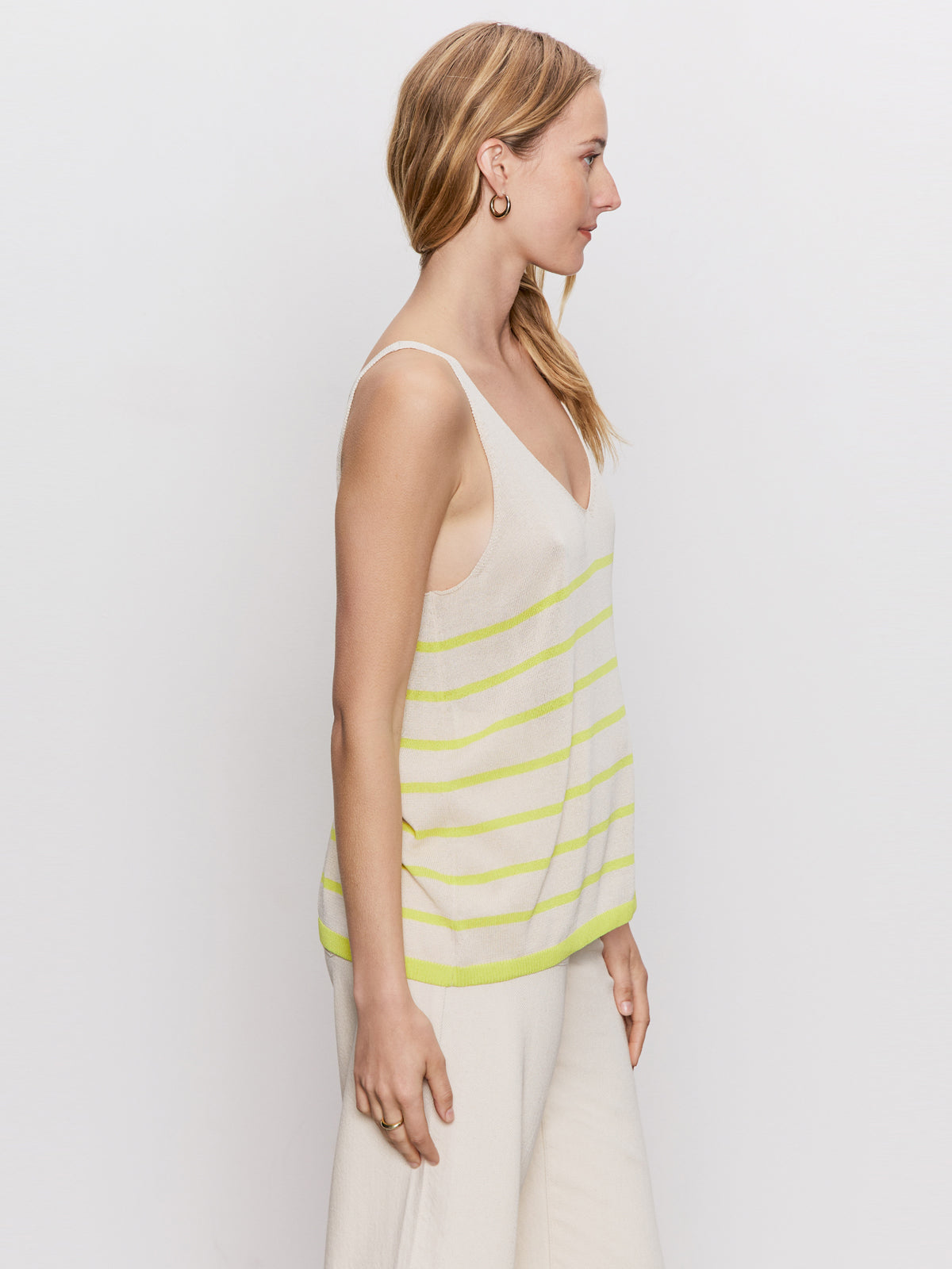 A woman with light brown hair stands in profile against a white background, wearing Sanctuary Clothing's v neck knitwear tank in muslin desert lime stripe and cream pants.