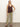 A woman poses against a plain light background in the Sanctuary Clothing V Neck Knitwear Tank Mesa, paired with light olive-green straight-leg pants and brown sandals. She has one hand in her pocket and a neutral expression.