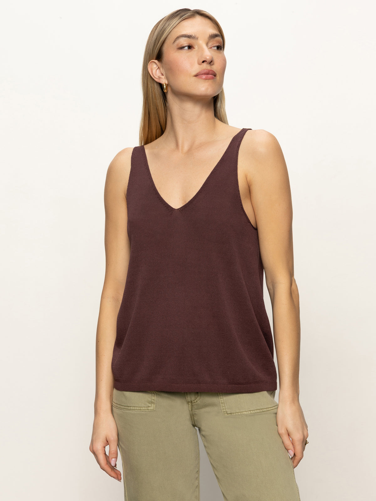 A woman with straight, light brown hair models the Sanctuary Clothing v neck knitwear tank mesa in dark brown, paired with light green pants. She stands against a plain background, gazing slightly to the side with a neutral expression.