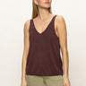 A woman with straight, light brown hair models the Sanctuary Clothing v neck knitwear tank mesa in dark brown, paired with light green pants. She stands against a plain background, gazing slightly to the side with a neutral expression.