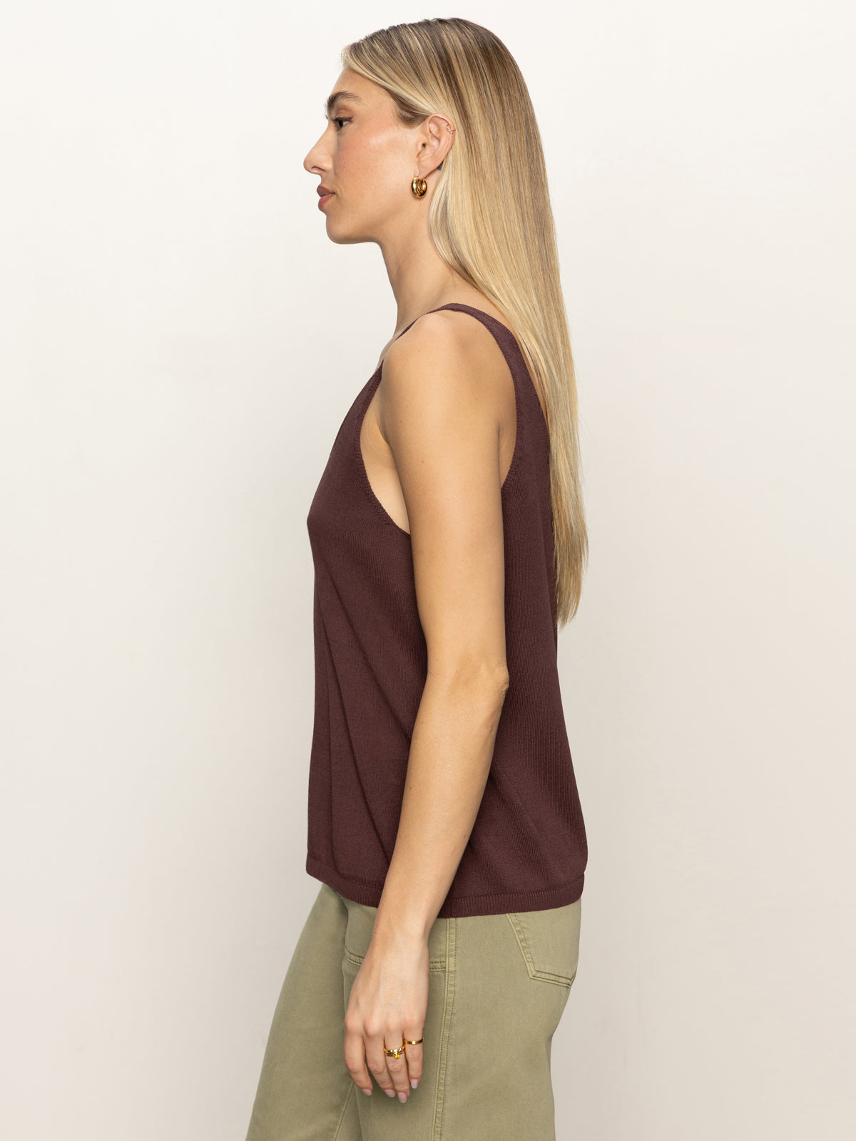 A woman with straight blond hair stands in profile against a plain light background, wearing the Sanctuary Clothing v neck knitwear tank mesa, a sleeveless dark brown top paired with light green pants.