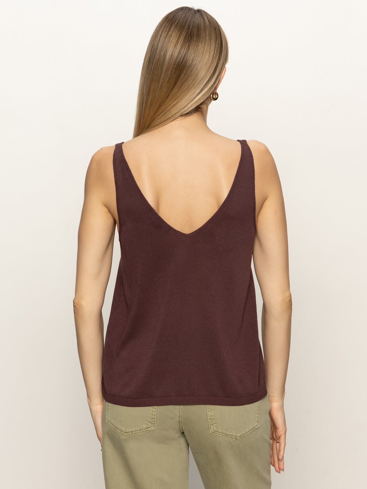 A woman with straight, light brown hair is seen from behind wearing the Sanctuary Clothing v neck knitwear tank mesa and light olive-green pants against a plain light background.